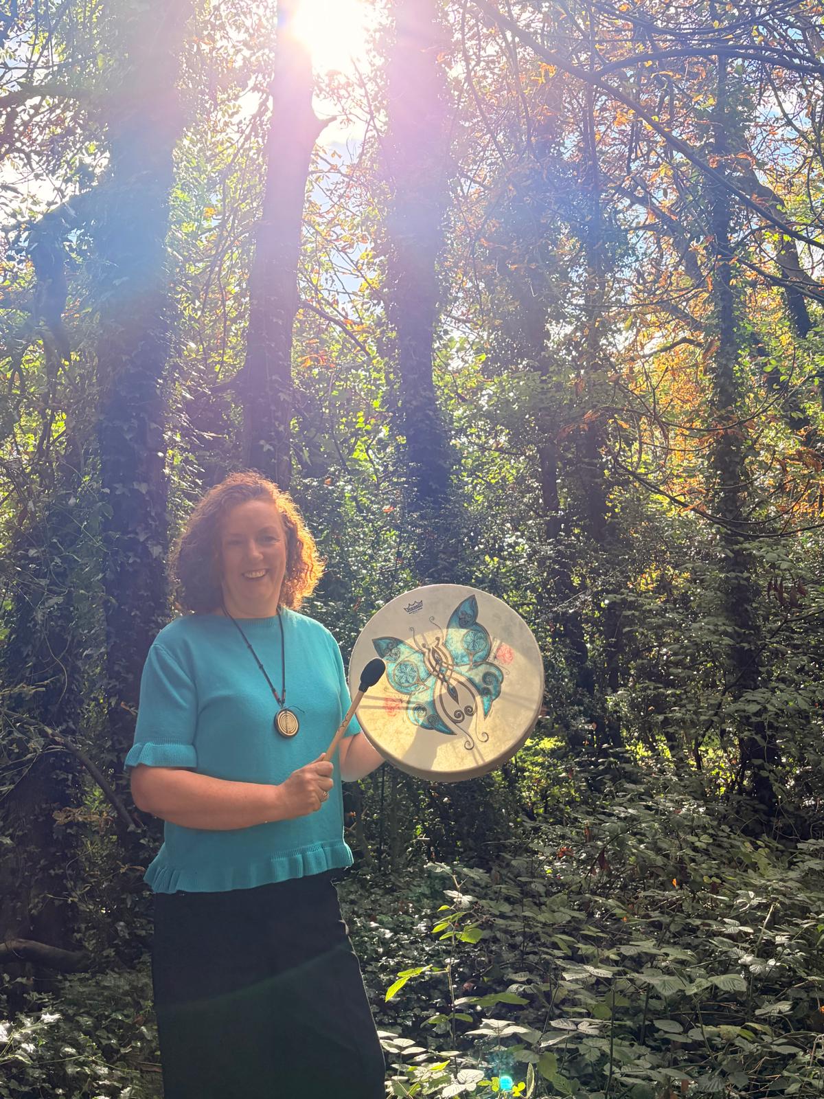 Deirdre O’Donnell smiling, holding a mallet beside a hand drum in woods
