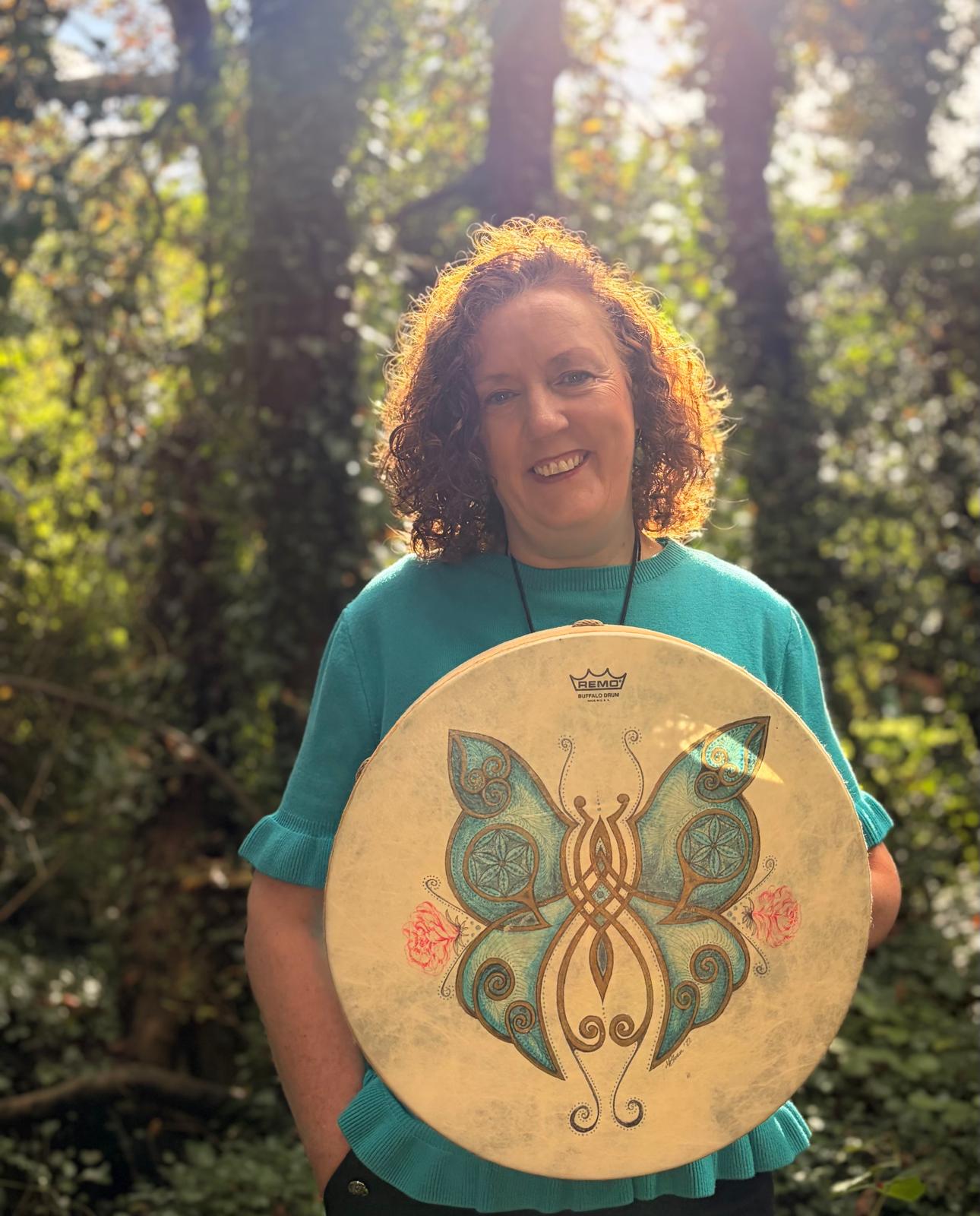 Deirdre O’Donnell smiling, holding a hand drum in sunlit woods
